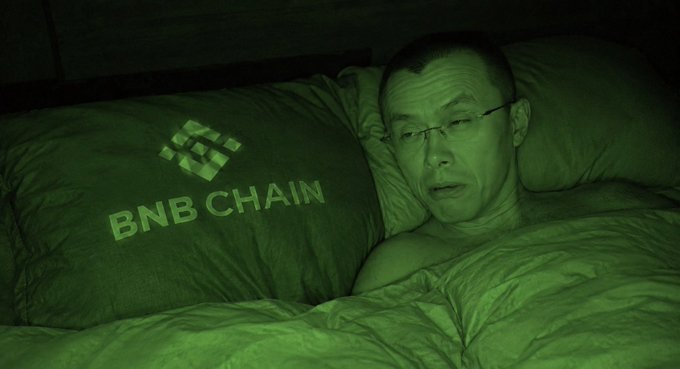 A man wearing glasses lies in bed next to a pillow featuring the BNB Chain logo, illuminated by a green night-vision effect.
