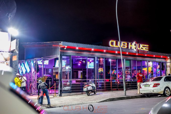 A nighttime exterior shot of a brightly lit nightclub called 'CLUB HOUSE OLYMPIC' with neon signage, people standing outside, and parked vehicles.