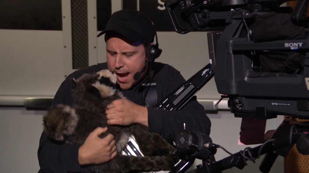 A behind-the-scenes photo showing a man wearing a cap and headset holding a raccoon while a large Sony professional video camera is positioned in the foreground.