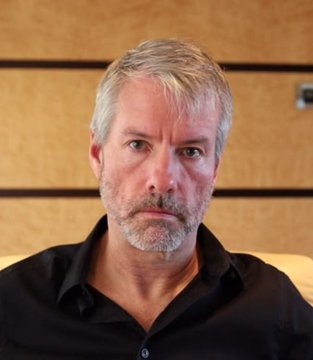 A portrait of a gray-haired man with a goatee wearing a black button-up shirt, seated on a beige couch.