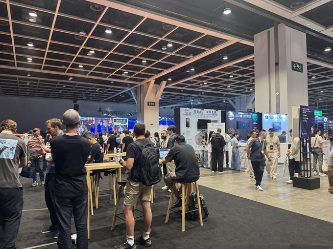 A crowded crypto/Web3 conference hall with attendees gathered around booths, including one for "Crypto Trading," under a large grid ceiling.