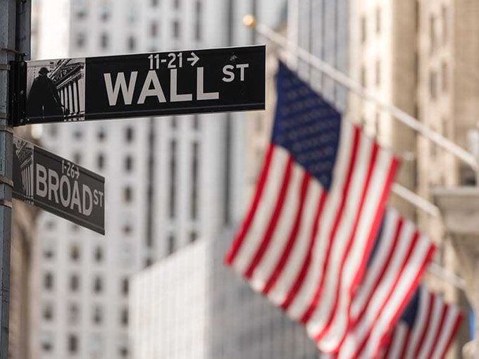 The image shows street signs at the iconic intersection of Wall St and Broad St in New York City's financial district, with American flags hanging in the background. This visual ties directly to the post's headline about $1 trillion in short-term debt flooding markets, as Wall Street—the symbolic heart of U.S. finance—warns of escalating borrowing costs.