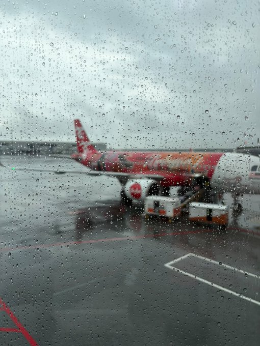 The image shows a plane parked at an airport with raindrops on its windows. There are two people visible near the airplane, possibly attending to some tasks or preparing for their next flight. The scene also includes a red line running across the bottom of the photo, which could be a runway or taxiway marking system.