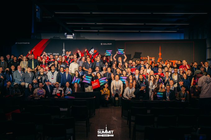 A large group of attendees poses together at the Solana Summit Canada 2026, holding colorful Solana-branded signs in front of a stage backdrop displaying "SOLANA," "SUPERTEAM," and city skyline graphics, with "THE SOLANA SUMMIT" logo visible at the bottom of the image.