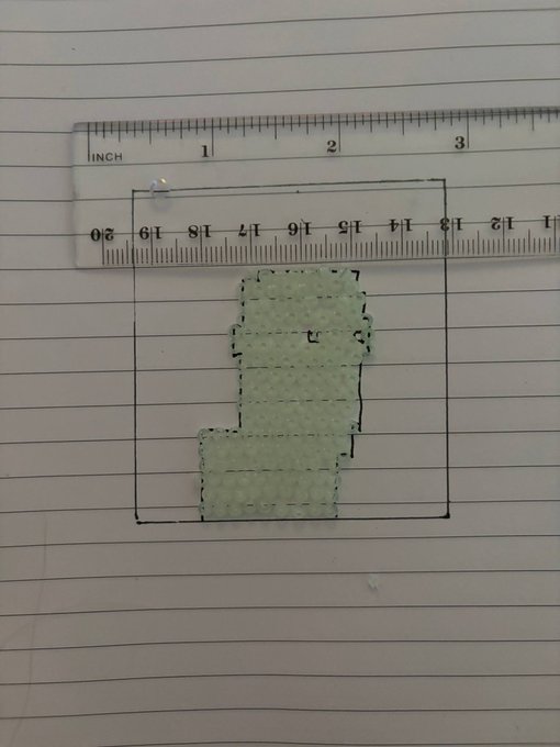 A hand-drawn diagram on lined notebook paper showing a figure (resembling an 'L' shape) drawn with a dashed outline inside a rectangular box, with a clear plastic ruler placed above showing inches and centimeters.