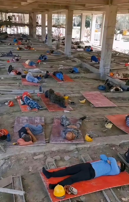 Construction workers resting on mats laid out across the floor of an unfinished building, many wearing hard hats placed beside them.