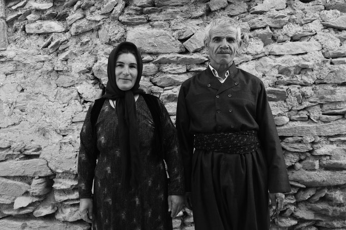 An elderly couple stands solemnly against a rough stone wall in this black and white portrait, with the woman wearing a headscarf and patterned dress, and the man in a traditional dark tunic with a sash.