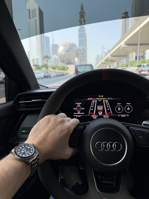 A driver's hand on an Audi steering wheel displays a digital dashboard, with a luxury watch visible on the wrist, while navigating a city street with modern skyscrapers.