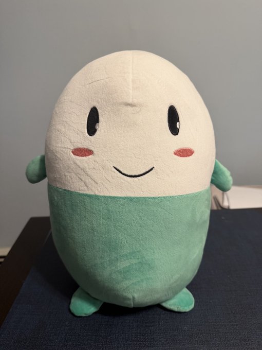 A smiling plushie with a cream-colored egg-shaped upper body, green lower half, tiny arms, and feet, sitting on a dark blue surface — matching the $Cupsey Upsey character plush from HappyPillco mentioned in the post.