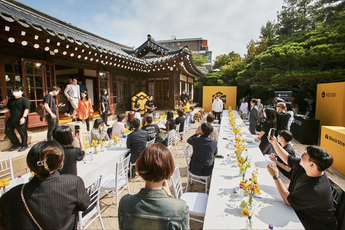 An outdoor event at a traditional Korean hanok building with long banquet tables, attended by guests in formal attire, with ASTER and BlockStore branding visible on yellow signage.