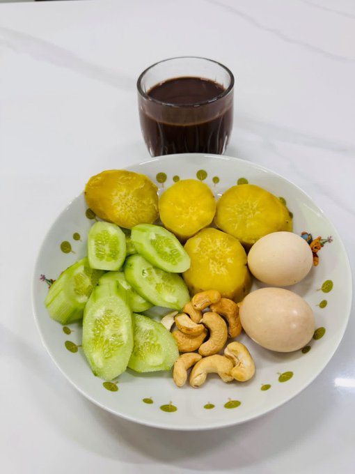 A plate containing yellow potatoes, sliced cucumbers, cashews, and two eggs, accompanied by a glass of dark beverage on a white surface.