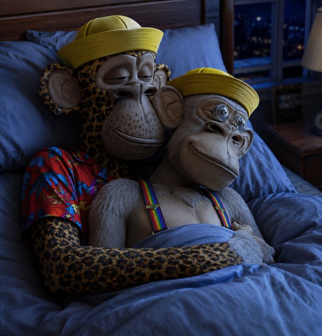 Two Bored Ape Yacht Club NFT characters, one wearing a yellow cap and leopard-print fur and the other in a yellow beanie with glasses and rainbow suspenders, snuggle together under blue sheets in bed, matching the cozy "goodnight" vibe of the tweet.