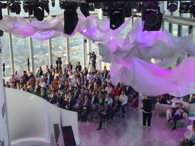 An audience of over 100 attendees fills a high-rise venue with floor-to-ceiling windows overlooking a cityscape, seated attentively under draped white fabric illuminated by purple stage lights and surrounded by professional lighting rigs—visually confirming the "standing room only" crowd at the Solana & Suits event.