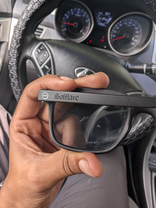 A hand holds a pair of black 'Solflare' sunglasses inside a car, with the steering wheel and dashboard gauges visible in the background.