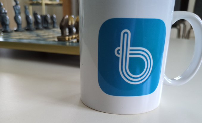 The image features a white mug with a blue logo that reads "b" on it, placed on a table next to a chess set. The background of the photo shows a wooden floor and a wall, providing context for the setting where the mug is located. The text in the image suggests that the mug has been used by @bitrefill for almost ten years, indicating its popularity among users.
