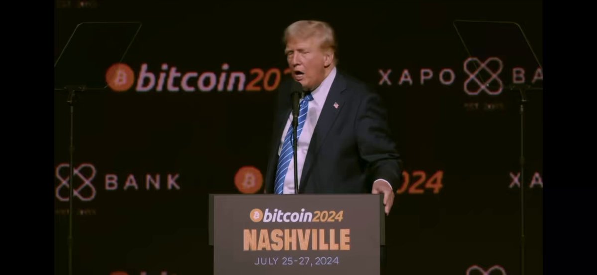 The image features a man standing at a podium, giving a speech or presentation. The background of the image shows a large screen displaying the text "BITCOIN2024 NASHVILLE JU 25, 2026 JUN 24, 2026". This suggests that the event is related to Bitcoin and might be taking place in Nashville. The man's attire includes a suit and tie, indicating a formal setting for the event.