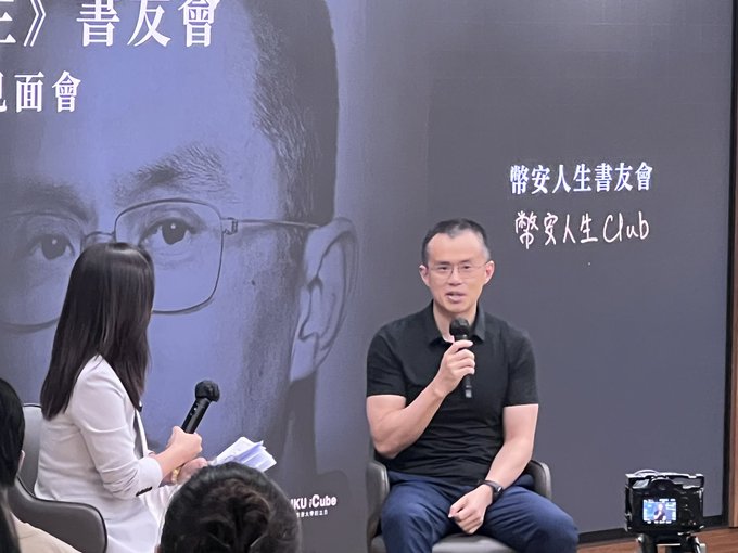 A man wearing glasses and a black polo shirt speaks into a microphone at a book club event, with a large backdrop reading "幣安人生書友會 / 幣安人生Club" featuring his face.