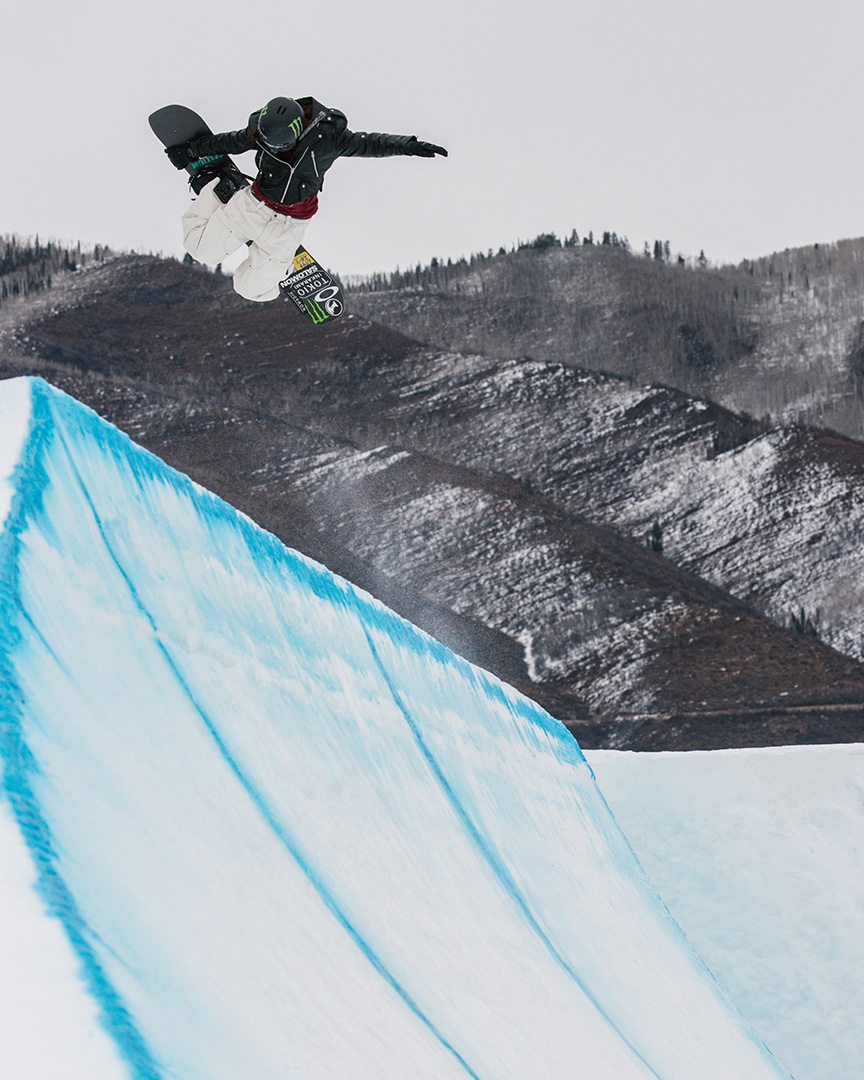 A snowboarder catches air above a halfpipe, performing a grab trick while soaring against a backdrop of snow-covered mountains, with "Monster" branding visible on the bottom of the snowboard.