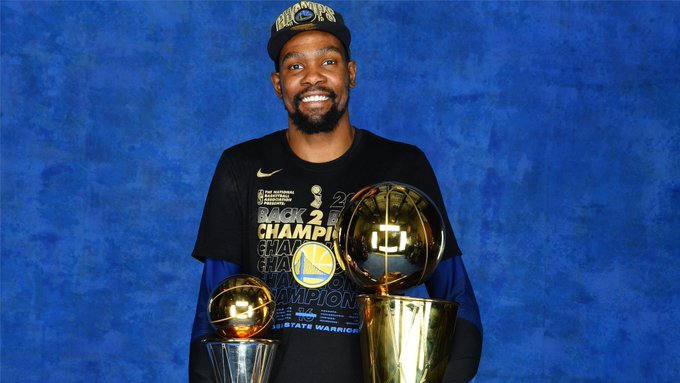 A smiling Kevin Durant poses with two NBA championship trophies and Finals MVP trophies while wearing a "Back 2 Back Champions" shirt and Warriors cap, celebrating the Golden State Warriors' 2017-2018 championship wins—an image tied to his off-court success, including his lucrative early investment in Hugging Face, which has reportedly grown to an estimated $60 million from Nvidia's $12.9 billion acquisition of the company.
