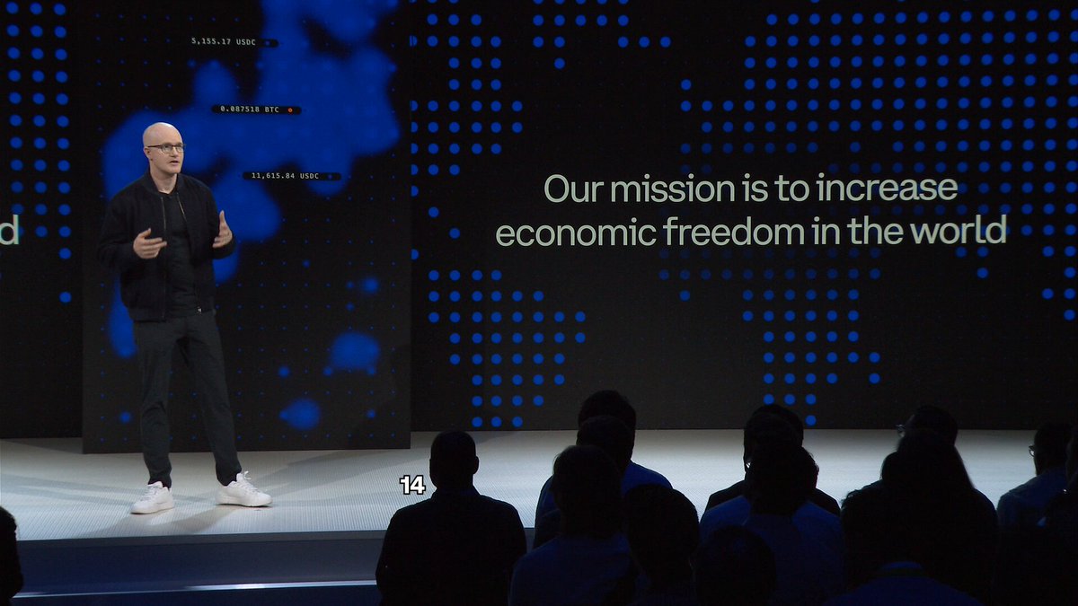 The image shows an event where a man is standing on stage, giving a speech or presentation. There are several people sitting in chairs around him, attentively listening to his words. The background of the scene features a large screen displaying information related to Bitcoin, possibly indicating that this event is about cryptocurrency or blockchain technology.