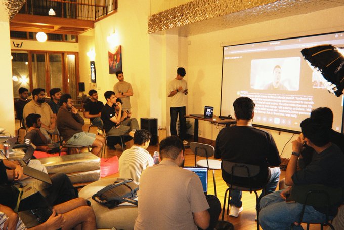 A group of people gather in a lounge for a viewing party, watching a presentation on a large projector screen.