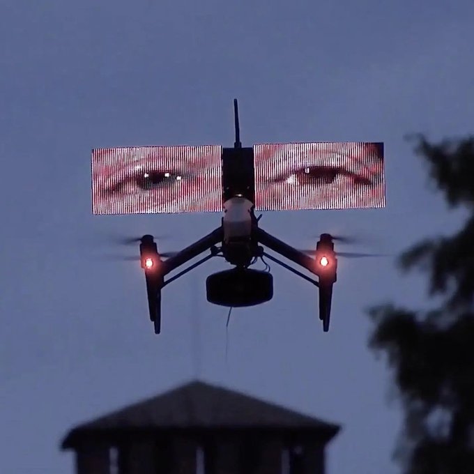 A drone with glowing red lights hovers against a blue sky, displaying a digital screen showing a close-up of eyes on its wings, above the silhouette of a gazebo-like structure.