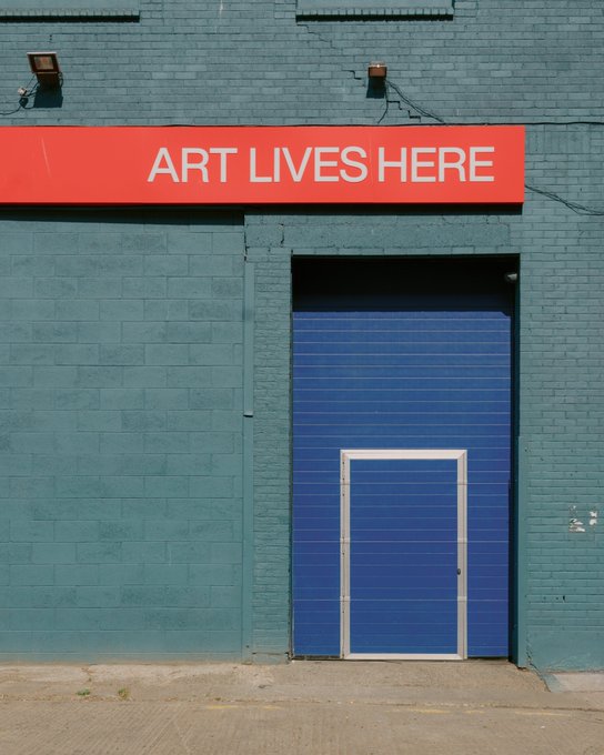 A blue brick building features a large blue garage door with a smaller pedestrian door, and a red banner above reading 'ART LIVES HERE'.