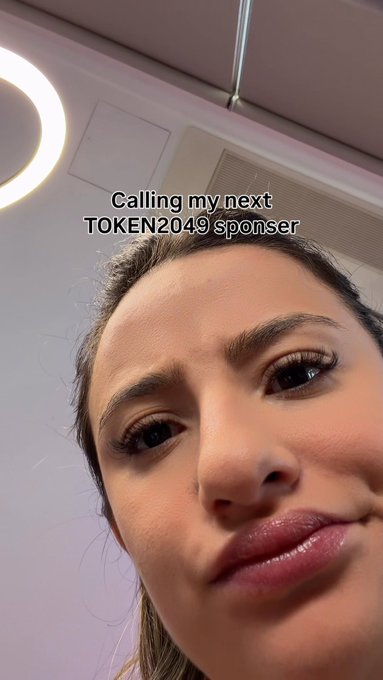 A close-up selfie of a young woman looking up at the camera with the text overlay 'Calling my next TOKEN2049 sponsor' displayed above her.