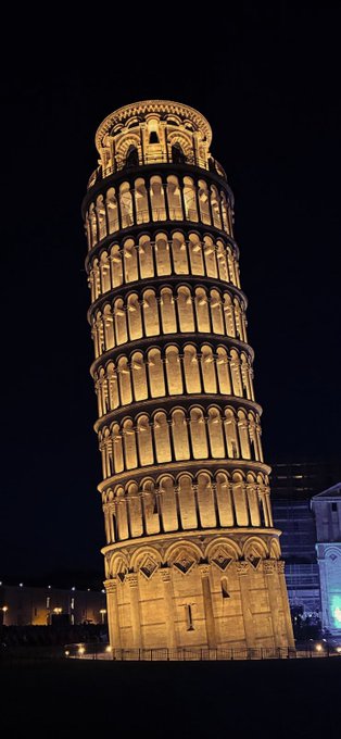 The Leaning Tower of Pisa illuminated in golden light against a black night sky, showing its iconic tilted cylindrical structure with arched colonnades.