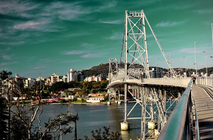 A scenic photograph of a large suspension bridge (appears to be the Hercílio Luz Bridge in Florianópolis, Brazil) over water with a cityscape and hills in the background.