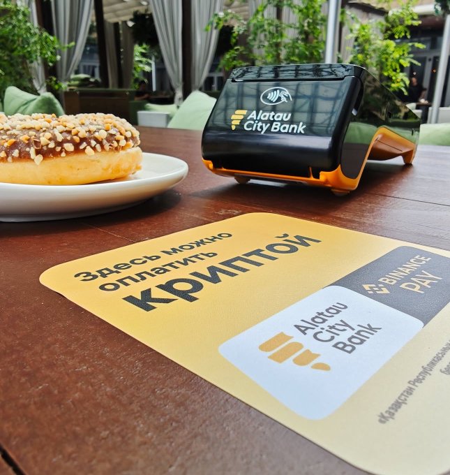 A Subway restaurant table with a donut and an Alatau City Bank payment terminal and QR card ('Здесь можно оплатить КРИПТОЙ').