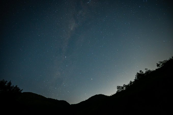 A night sky filled with countless stars displays the Milky Way stretching vertically across the frame, with the dark silhouettes of hills and trees visible along the bottom edge.