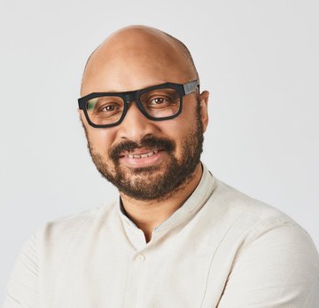 A professional headshot of a bald man with a beard and dark-rimmed glasses, wearing a cream-colored shirt against a white background.
