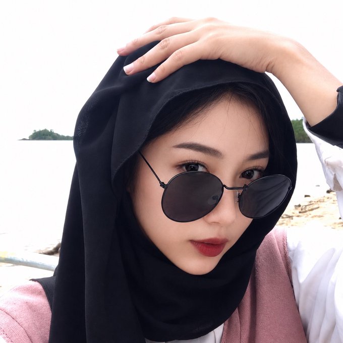 A selfie of a woman wearing a black hijab, round black sunglasses, and red lipstick, posing near a beach with her hand on her head.
