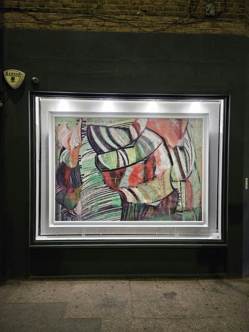 A framed abstract painting illuminated by an interior light box, displayed against a dark wall at night, featuring swirling shapes in red, green, and black, with a 'BANKAM' security sticker visible on the wall to the left.
