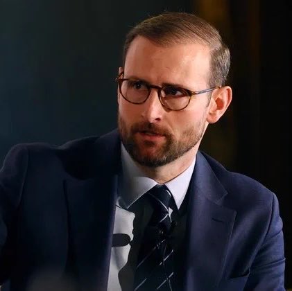 A close-up portrait of a bearded man wearing glasses and a dark suit with a striped tie, appearing to be in a professional interview or panel setting.