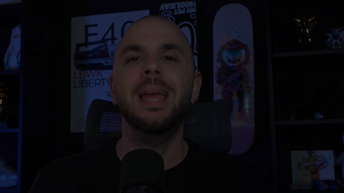 The image shows a man wearing a black shirt sitting behind a microphone, smiling at the camera while talking into it. The background features a wall with various posters and decorations, including one that reads "E4LVA LIBERTY". There is also an image of a skateboard on the wall next to the poster.