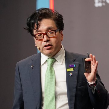 Tom Lee, wearing red-framed glasses and a green tie, speaks while holding up a smartphone during a presentation, illustrating his latest commentary that the Federal Reserve "chose violence" with its rate hike, which he views as setting up an upcoming equity rally.