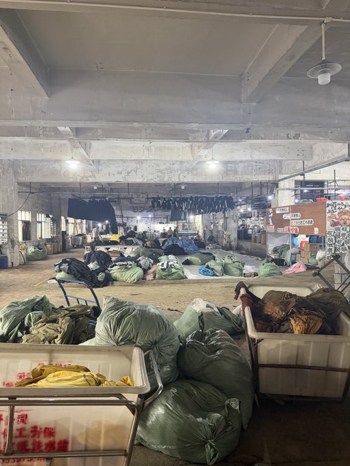 A warehouse interior filled with large green and yellow sacks of laundry, with workers sorting items and clothing hanging from ceiling racks.