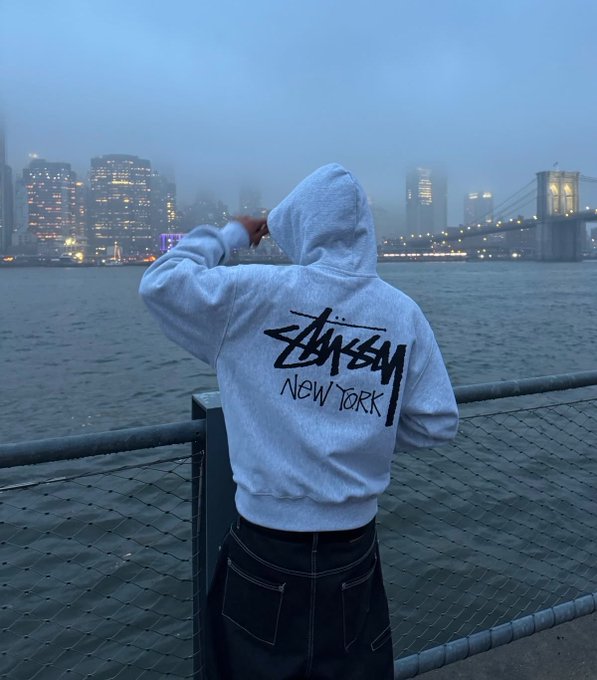 A person wearing a light grey Stüssy 'New York' hoodie stands with their back to the camera, looking out over a body of water towards a foggy city skyline featuring the Brooklyn Bridge.