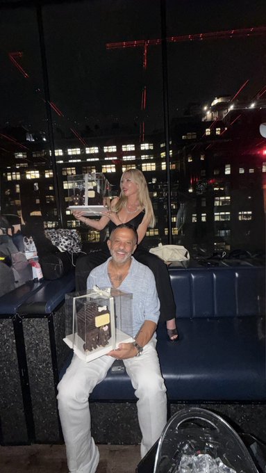 A man with a gray beard sits on a blue couch holding a clear display case containing a chocolate structure, while a blonde woman stands behind him holding another display case.