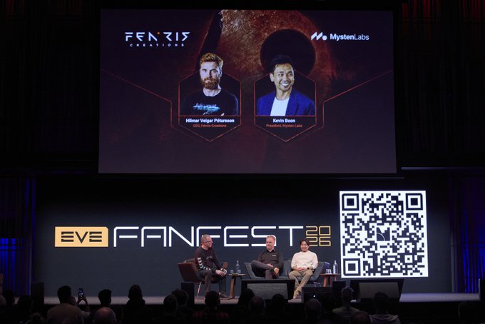 The image shows a stage with two men sitting behind a table, likely participating in a panel discussion or interview. The audience is seated in front of them, watching the event unfold. A large screen displays the names of the panelists, including "Sui Network" and "Fenris", as well as other relevant information about the event.