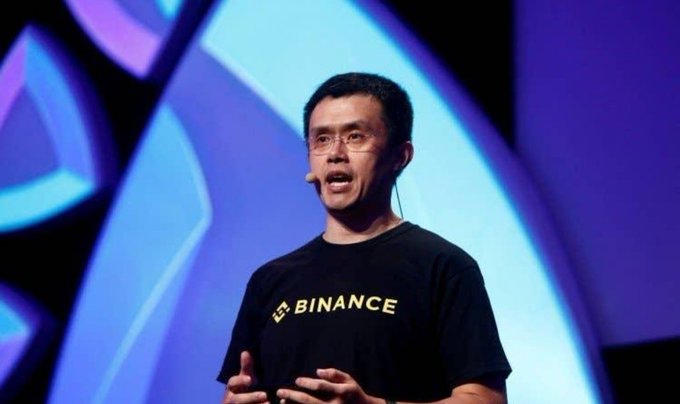 A man wearing a black "BINANCE" t-shirt speaks on a stage with a headset microphone, gesturing with his hands against a blue-toned backdrop. The image aligns with the post's theme about crypto's growing role in the global financial system, with Binance being one of the world's largest cryptocurrency exchanges.