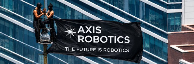 Two workers on a cherry picker hanging a large black banner that reads 'AXIS ROBOTICS - THE FUTURE IS ROBOTICS' against a modern glass building.