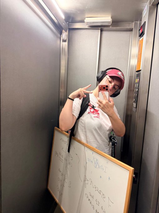 A person wearing a maroon Bored Ape Yacht Club cap takes a mirror selfie in an elevator while holding a whiteboard signed with messages like "Ape" and "BAYC," tying directly to the "Ladies of BAYC" theme by showcasing community participation in the NFT collection.