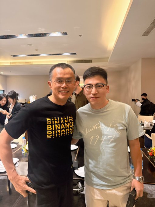 Two men pose together at what appears to be an EASY Residency graduation ceremony event, with the man on the left wearing a black "BINANCE" t-shirt and the man on the right wearing a light-colored "1947" t-shirt and glasses. The caption's reference to reuniting with @cz_binance (Changpeng Zhao) aligns with the Binance-branded shirt worn by the man on the left, suggesting this is the in-person meeting being celebrated.