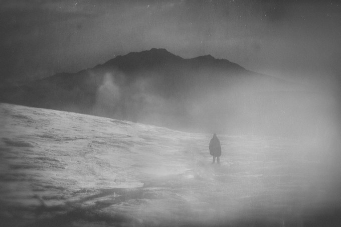 A moody black-and-white photograph of a lone figure walking across a misty, barren landscape with a mountain looming in the background, evoking a haunting, atmospheric mood.