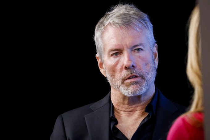 A close-up portrait of a gray-haired, bearded middle-aged man in a black blazer speaking against a dark background.