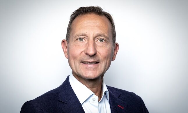 A professional headshot of a middle-aged man with short brown hair wearing a dark blazer over a light blue shirt, posed against a plain light background.