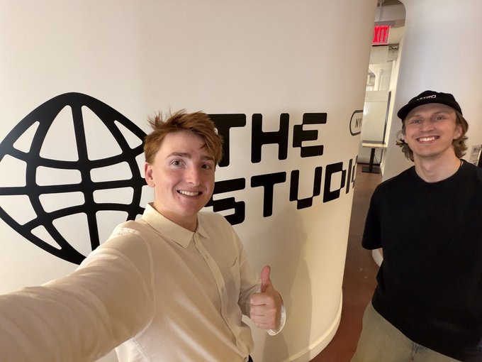 Two young men taking a selfie in front of a wall with "THE STUDIO" and a globe logo; one wears a white shirt and gives a thumbs-up, while the other wears a black cap with "LIVMO" and a black t-shirt.